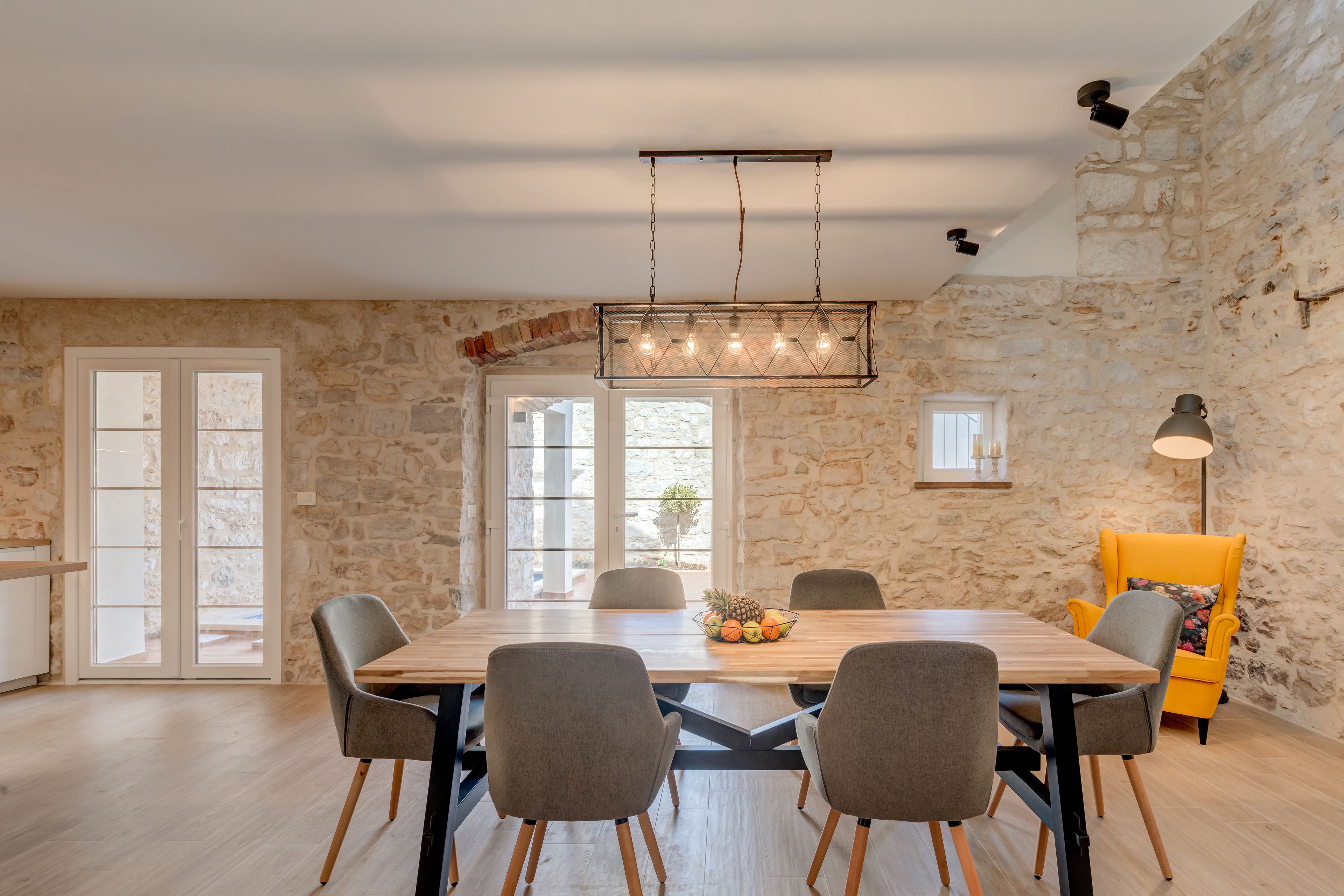 Un comedor con una mesa de madera, sillas grises, un frutero y una pared de piedra.