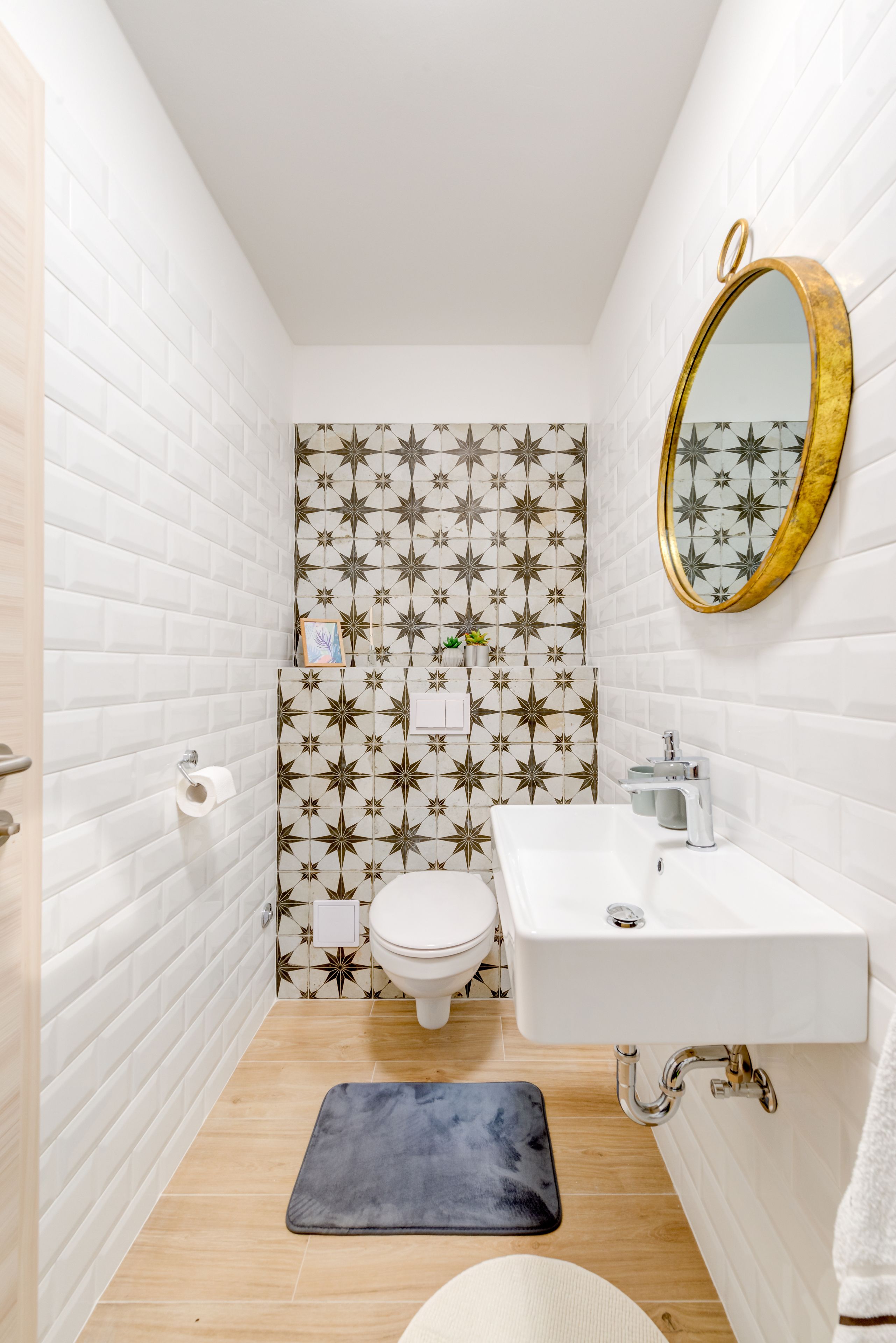 A compact bathroom with white subway tiles, a star-patterned accent wall, a white toilet, a white sink with a gold mirror, and a dark blue bath mat.