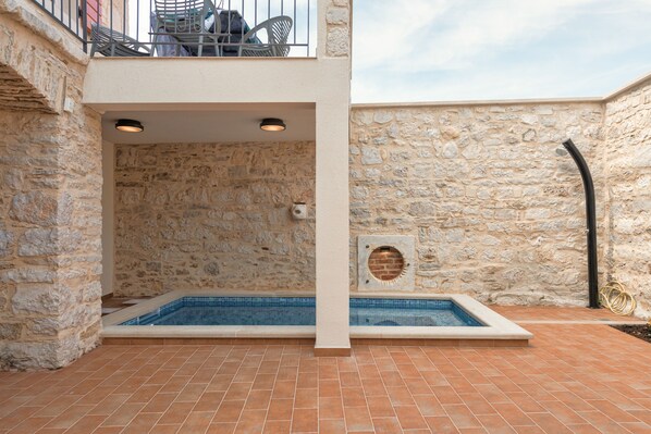 Zona de piscina al aire libre con paredes de piedra, suelo de baldosas, un espacio de descanso con sillas y un balcón con baranda.