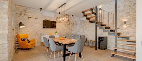 Un espace repas moderne avec une table en bois, des chaises grises et un fauteuil jaune. Un escalier avec une rampe en métal mène à un niveau supérieur.