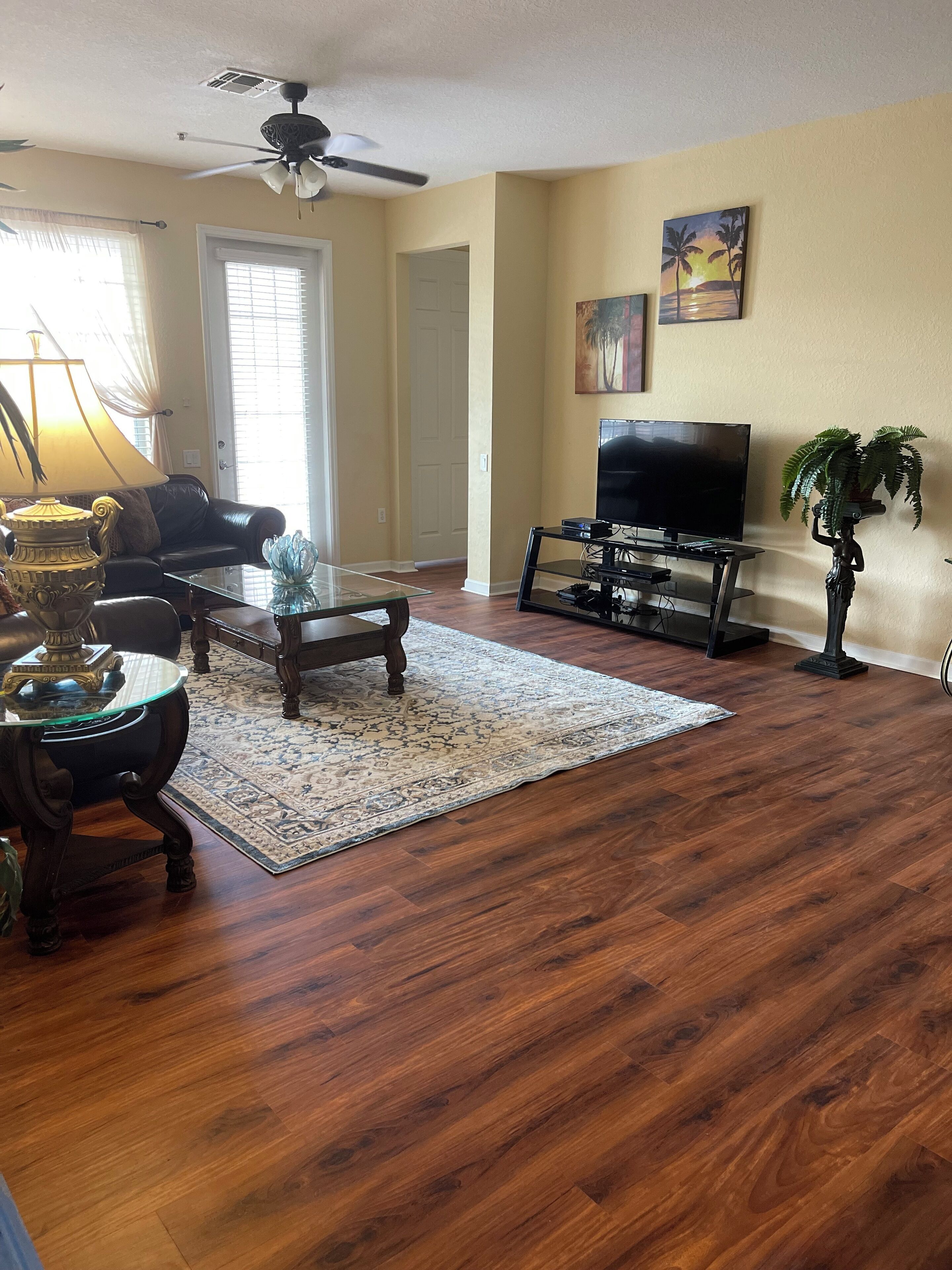 A living room with hardwood flooring, a ceiling fan, a TV, a coffee table, a sofa, and a decorative lamp.