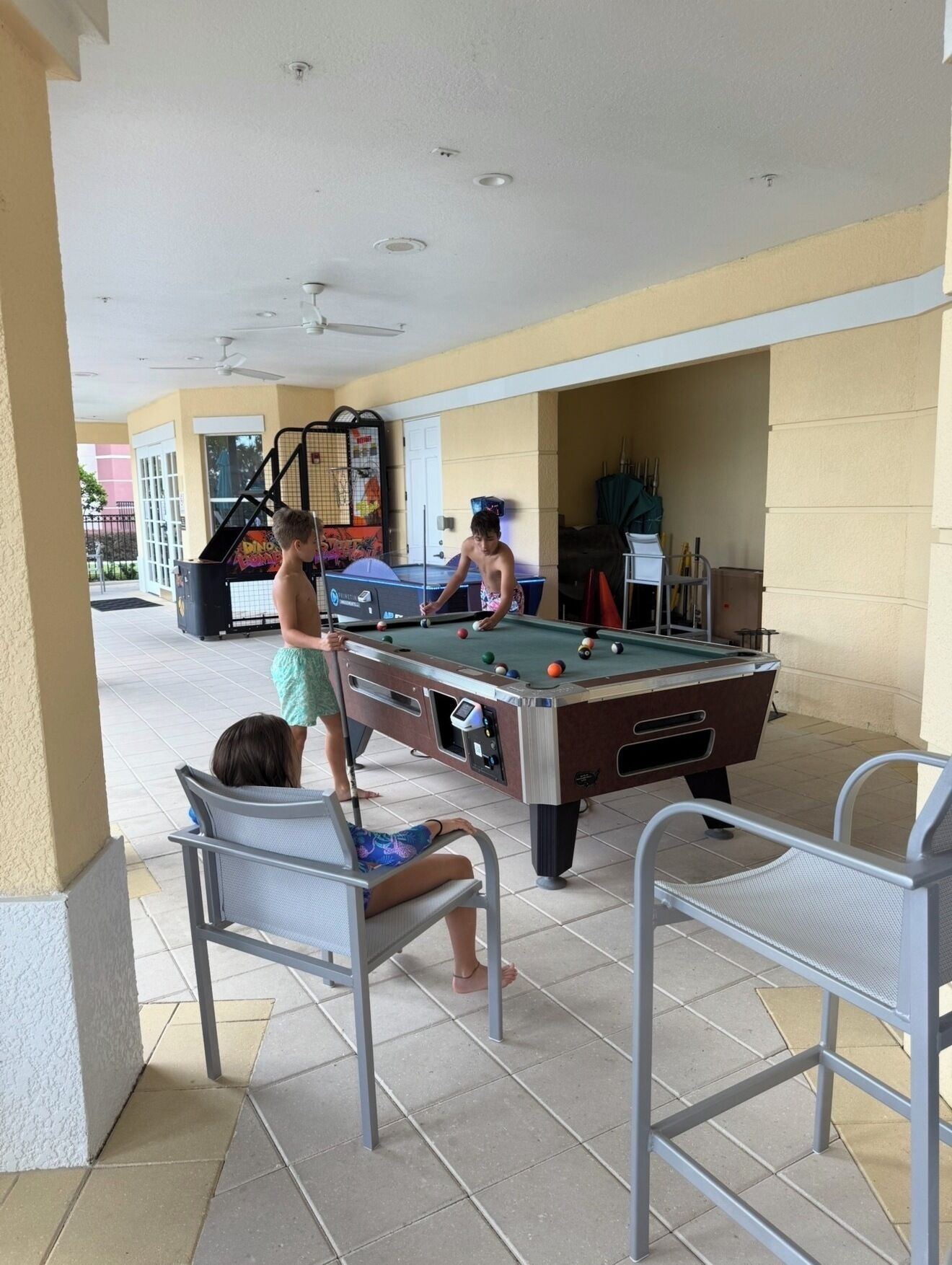 A pool table in an outdoor area with chairs and a person playing.