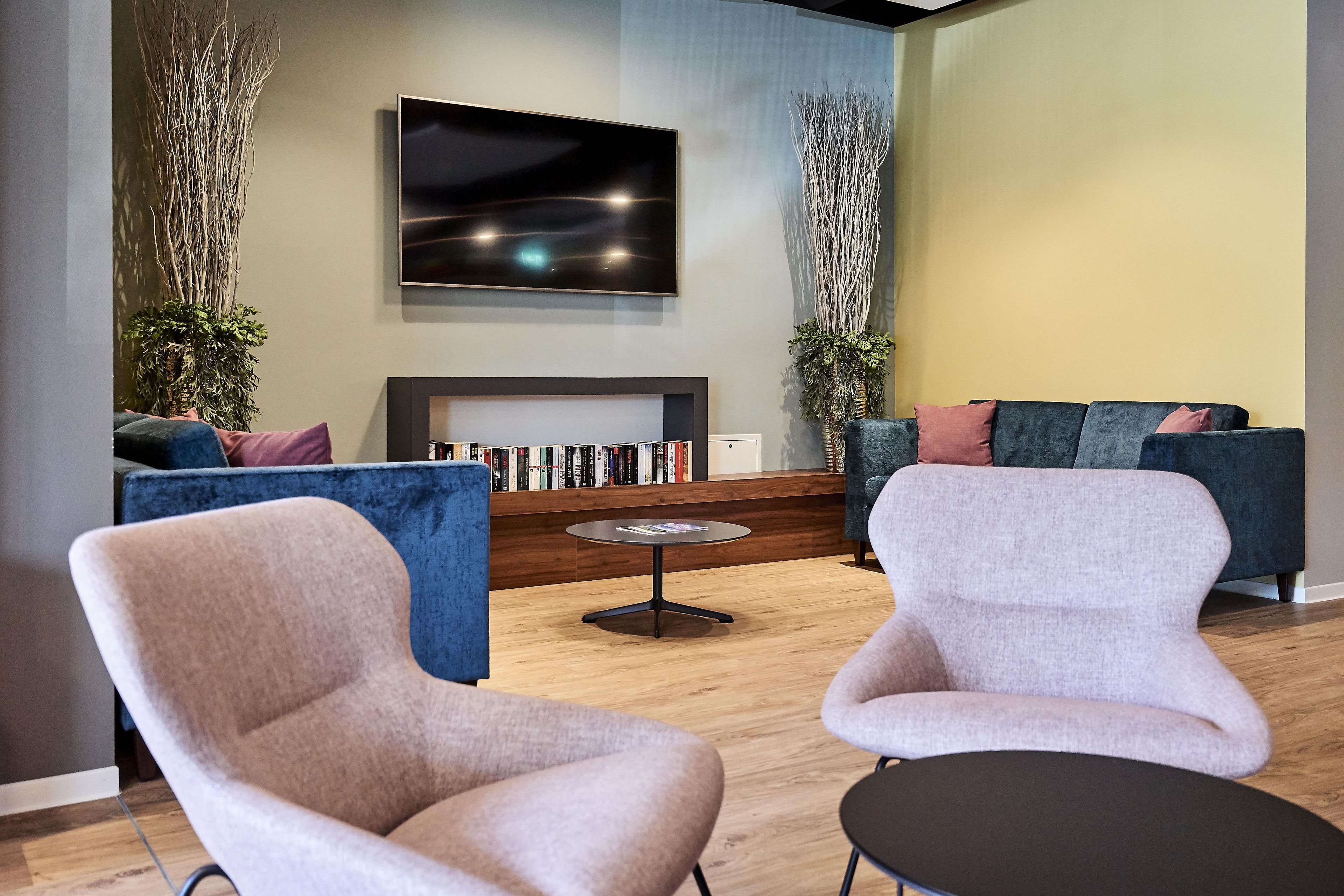 A modern living room with two armchairs, a sofa, a flat-screen TV mounted on the wall, and a bookshelf with books.
