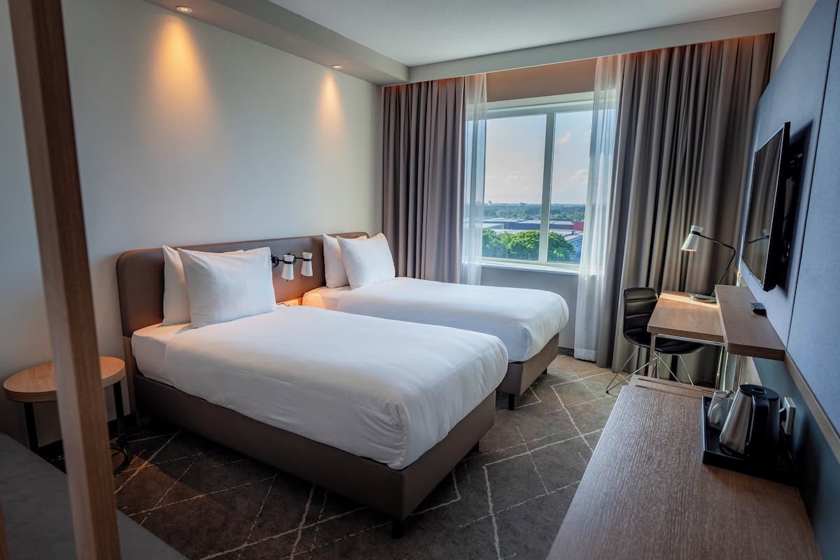 A hotel room with two beds, a desk, a chair, a TV, and a window with curtains.