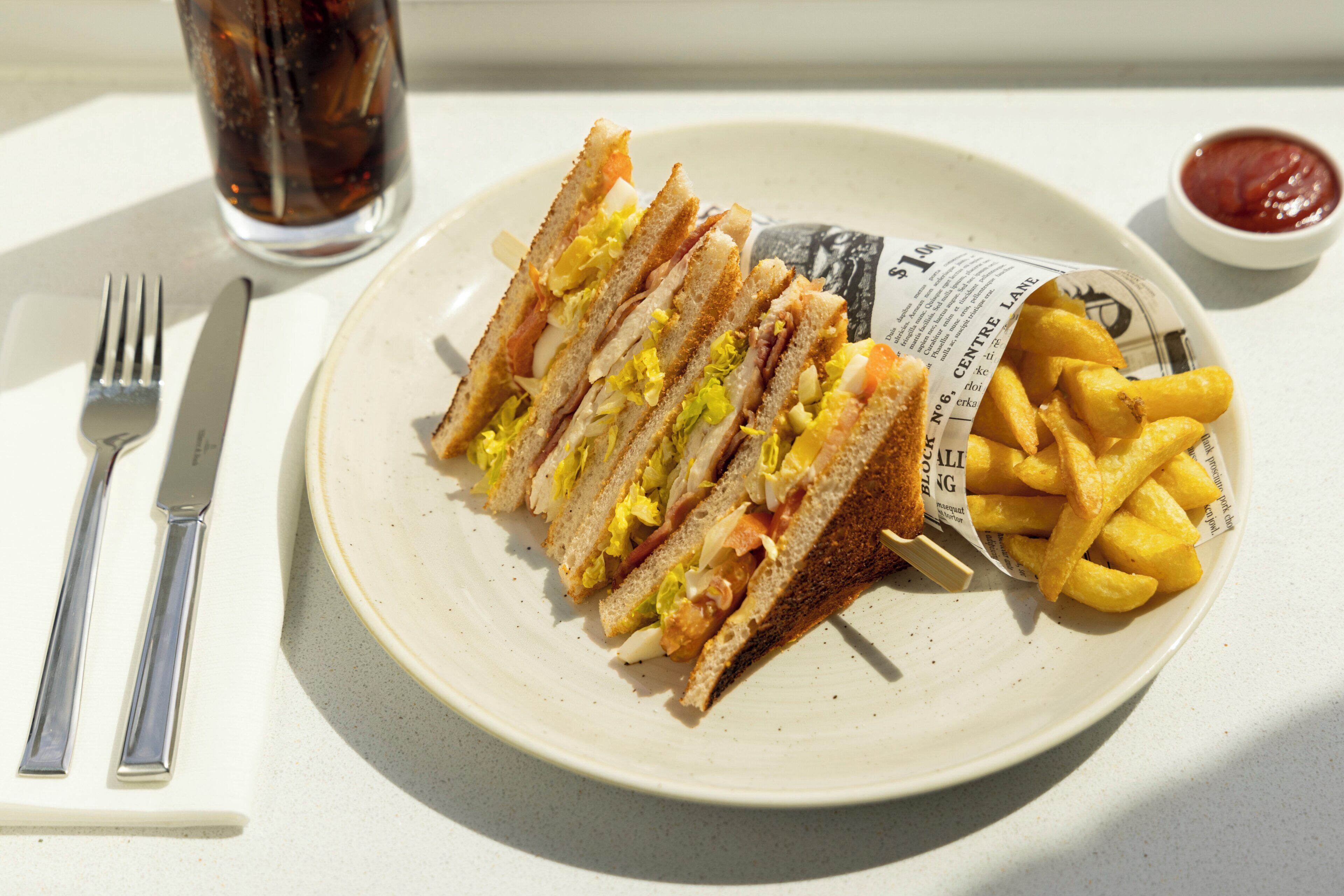 Un plato con un sándwich y papas fritas, un tenedor, un cuchillo y un vaso de refresco.