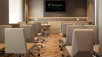 A hotel conference room with multiple chairs, a flat-screen TV displaying the Holiday Inn logo, and a carpeted floor.