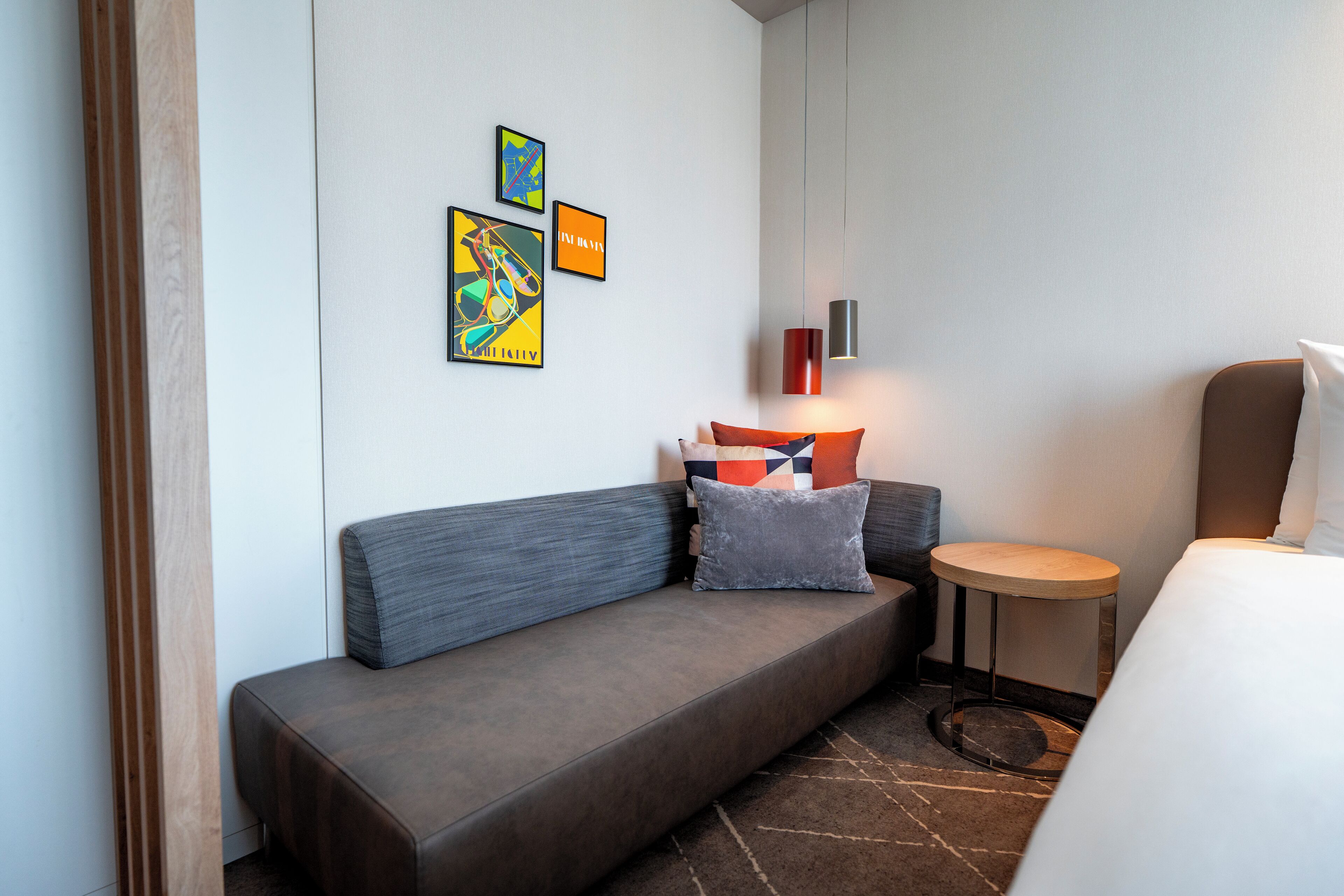 A modern hotel room with a grey sofa, a small round table, and a bed. There are three framed artworks on the wall.