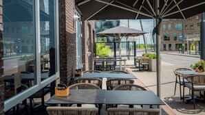 An outdoor seating area with tables and wicker chairs, large umbrellas, and a view of the street and buildings. - Holiday Inn Eindhoven Airport by IHG (Eindhoven)