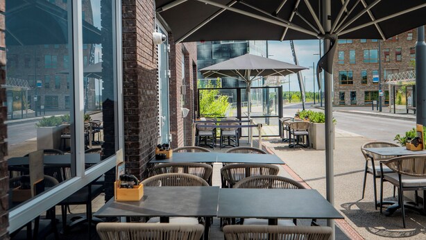 An outdoor seating area with tables and wicker chairs, large umbrellas, and a view of the street and buildings. - Holiday Inn Eindhoven Airport by IHG (Eindhoven)