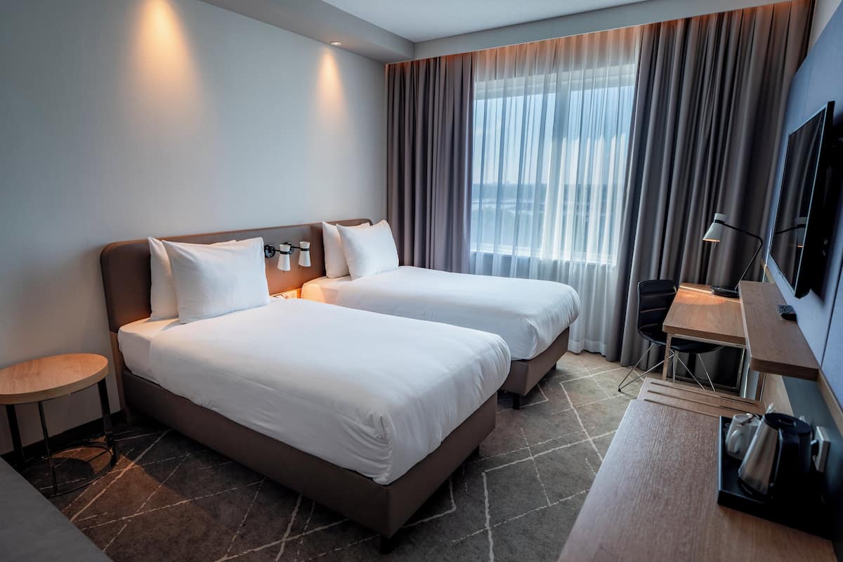 A hotel room with two beds, a desk, a chair, a TV, and a window with curtains.