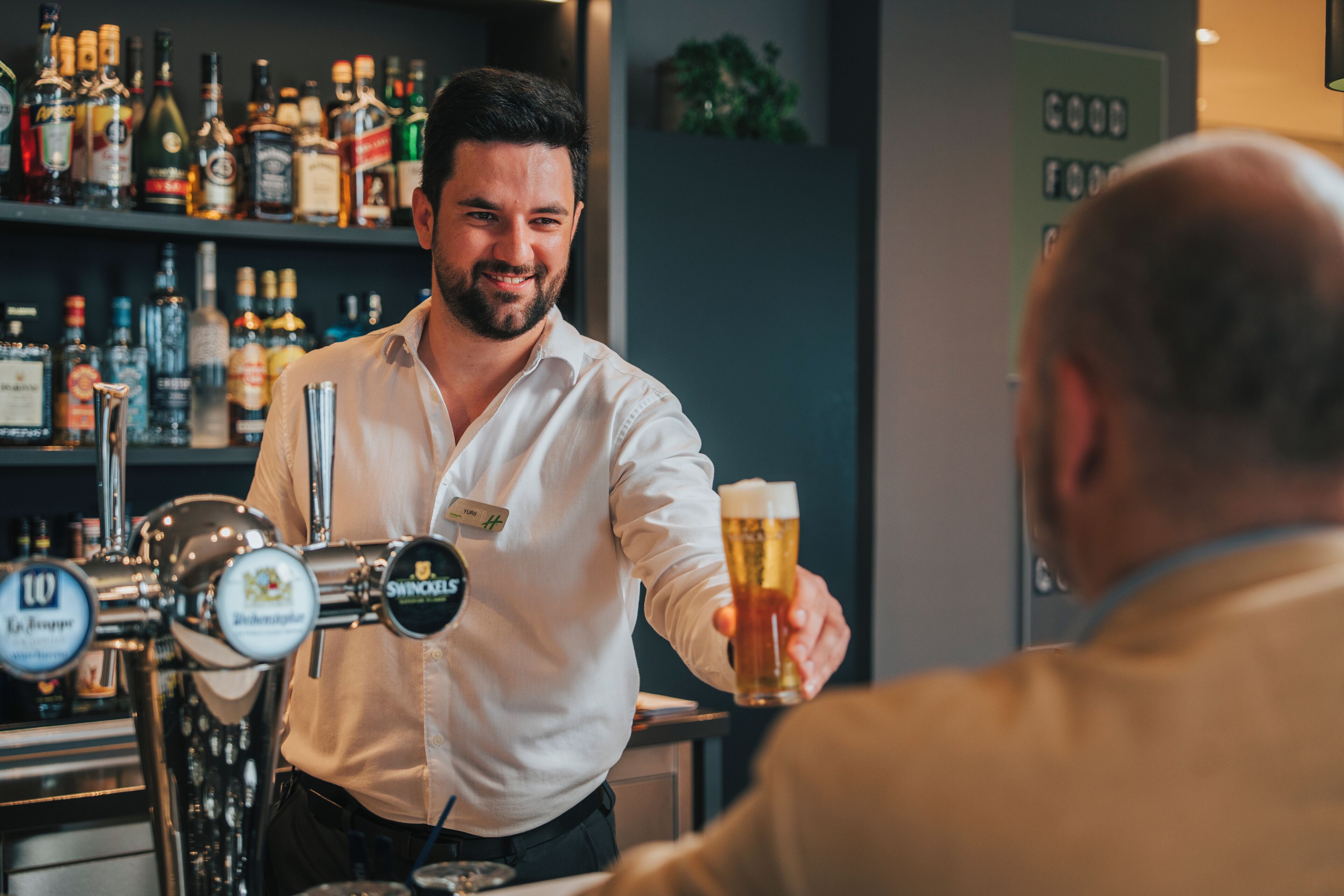 A bartender serving a beer to a customer in a bar with shelves of liquor bottles in the background.