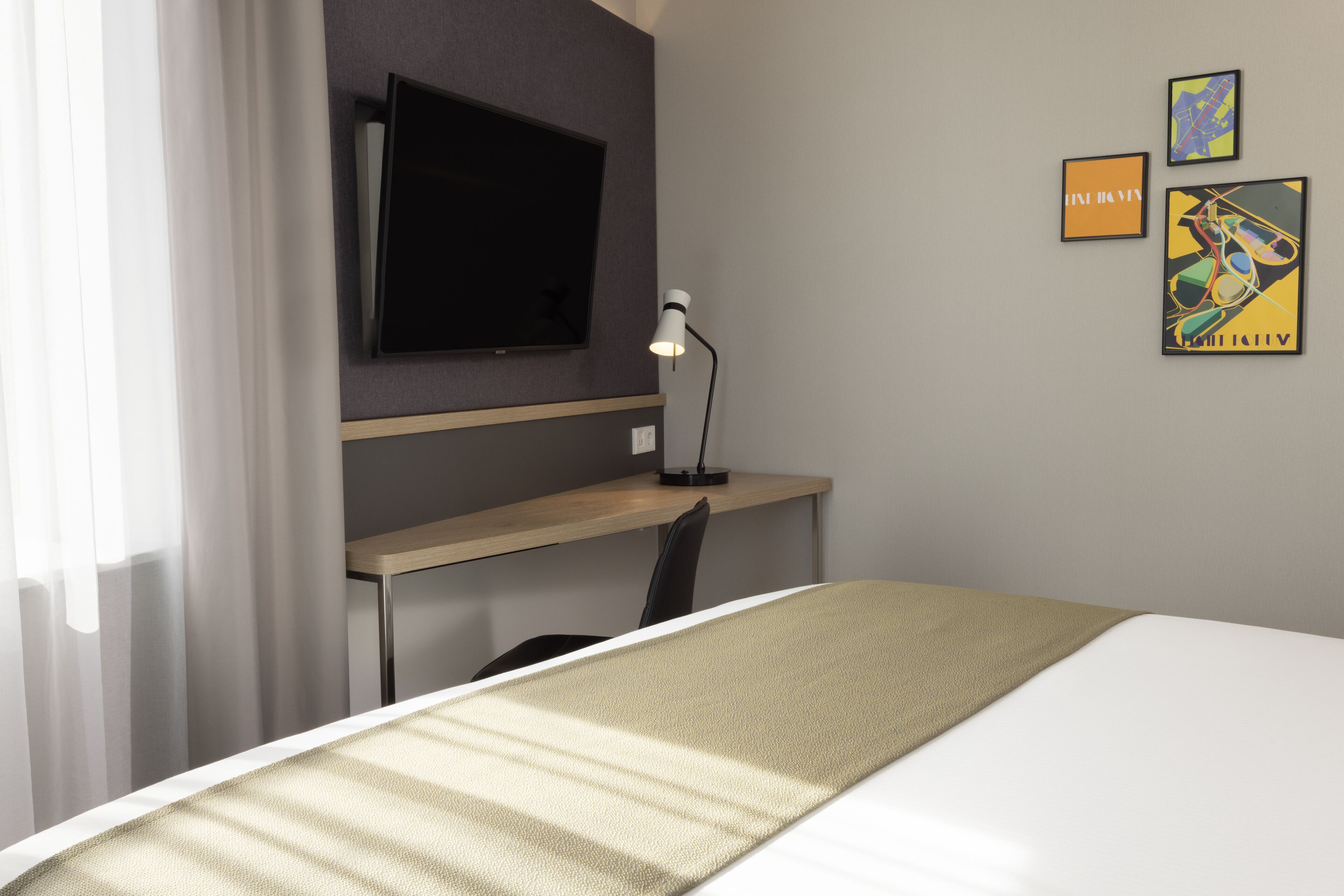 Habitación de hotel con cama, escritorio, silla, televisor montado en la pared y tres cuadros enmarcados.
