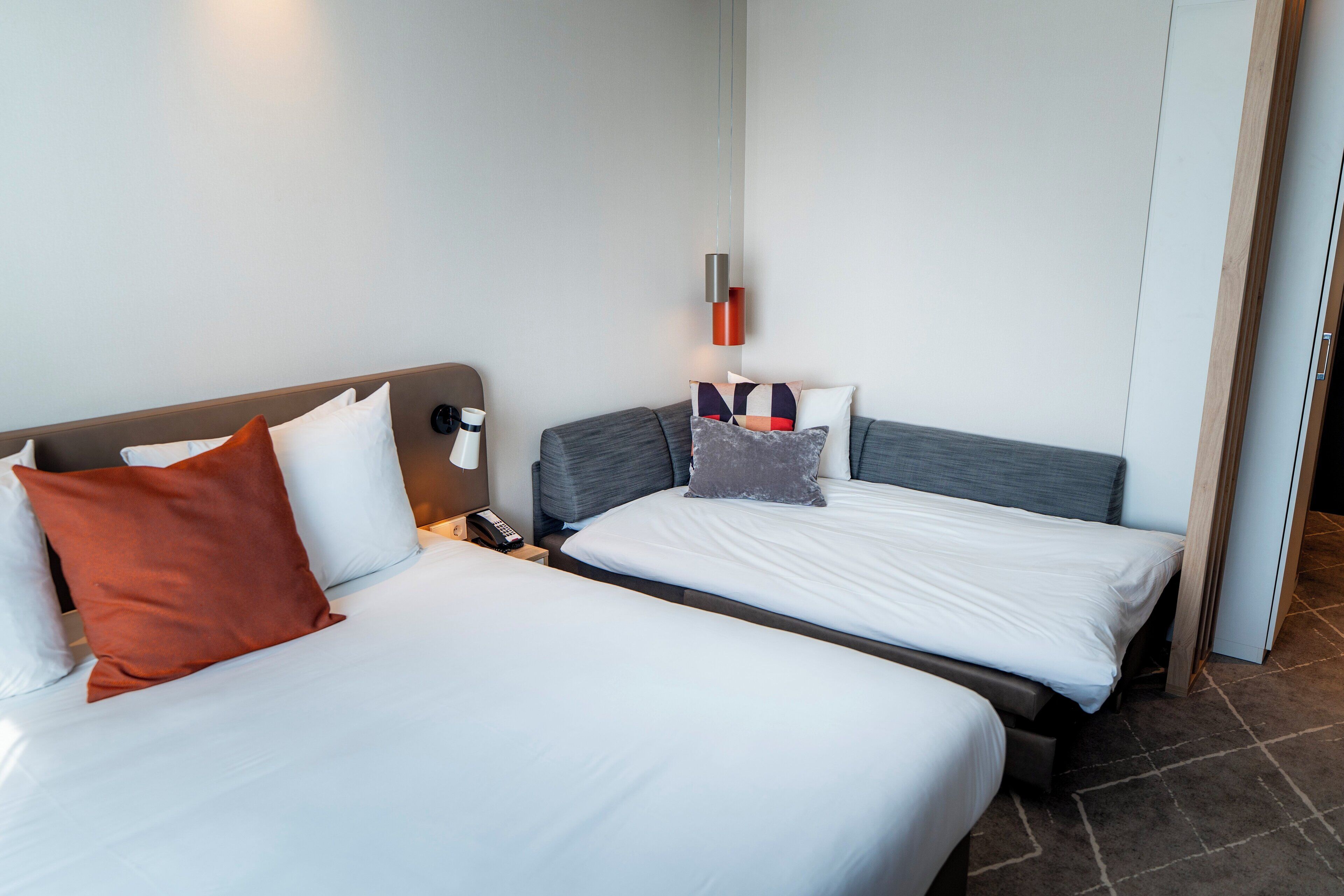Habitación de hotel con dos camas, cada una con ropa de cama y almohadas blancas, una mesita de noche con lámpara, y un aplique de pared.