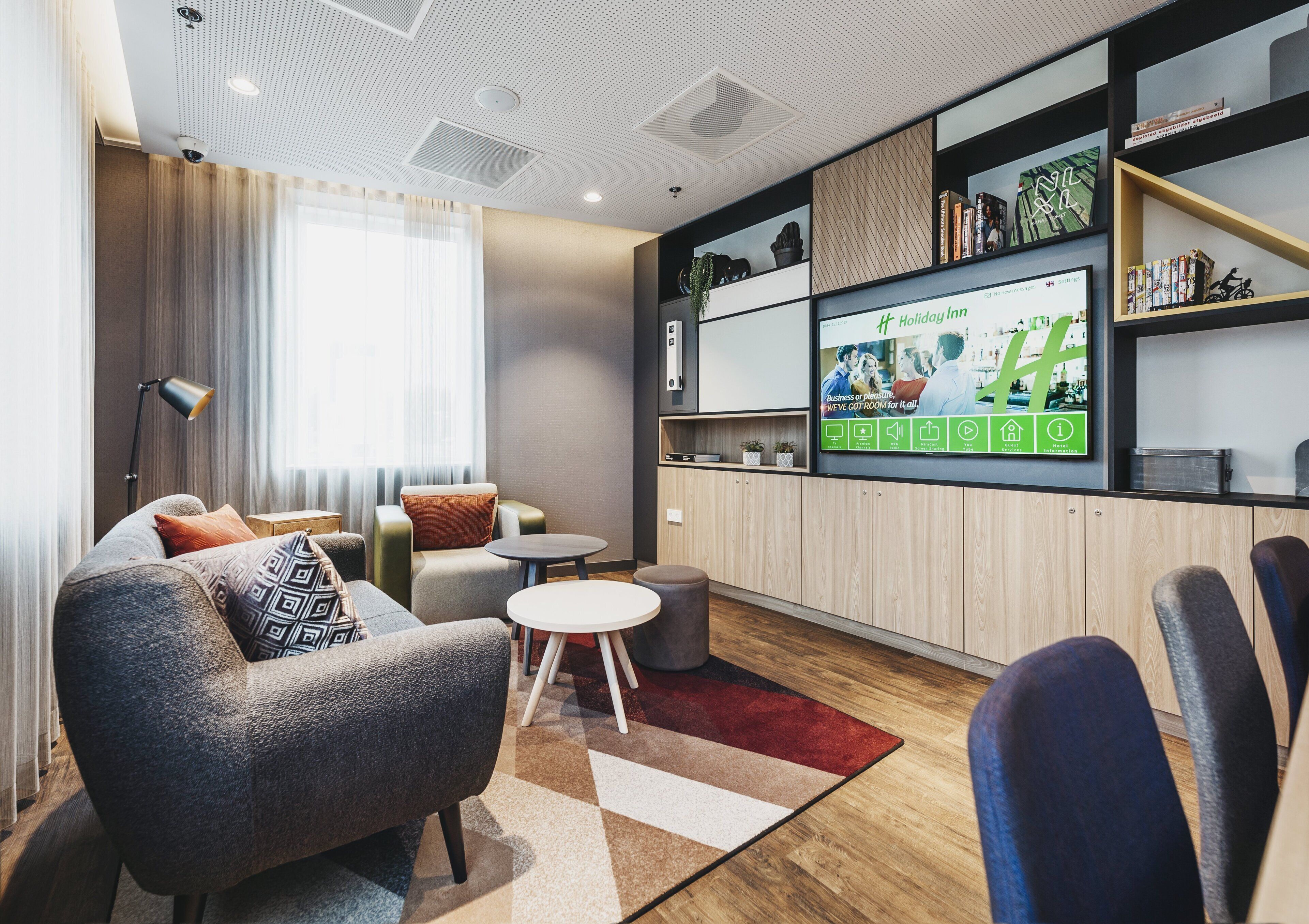Ein modernes Wohnzimmer mit einer Couch, Sesseln, einem Couchtisch und einem an der Wand montierten Fernseher, der eine Holiday Inn Werbung zeigt.