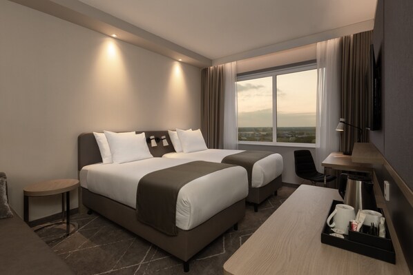 A modern hotel room with two beds, a desk, a chair, a coffee maker, and a view of the city. - Holiday Inn Eindhoven Airport by IHG (Eindhoven)