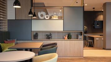 Un intérieur moderne avec une table à manger en bois, des chaises rembourrées, des luminaires suspendus et un coin cuisine doté d’une étagère intégrée où sont exposés des objets décoratifs.
