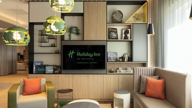 A modern hotel lobby with a large flat-screen TV, comfortable seating, and a bookshelf with books and decorative items. - Holiday Inn Eindhoven Airport by IHG (Eindhoven)