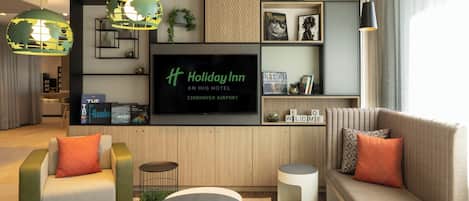 Un moderno vestíbulo de hotel con un televisor de pantalla plana grande, asientos cómodos y una estantería con libros y objetos decorativos.