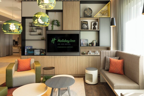 A modern hotel lobby with a large flat-screen TV, comfortable seating, and a bookshelf with books and decorative items. - Holiday Inn Eindhoven Airport by IHG (Eindhoven)