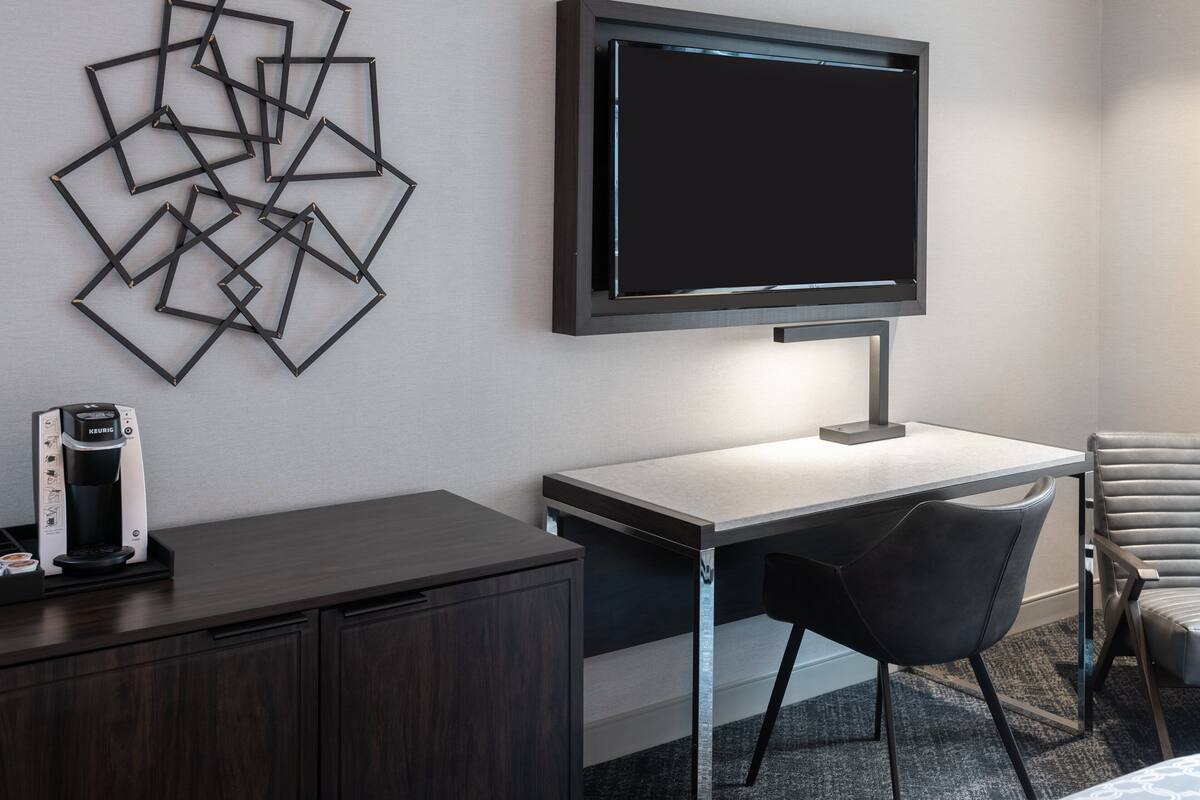 A modern hotel room with a flat-screen TV mounted on the wall, a desk with a chair, a coffee maker, and a decorative wall art piece.