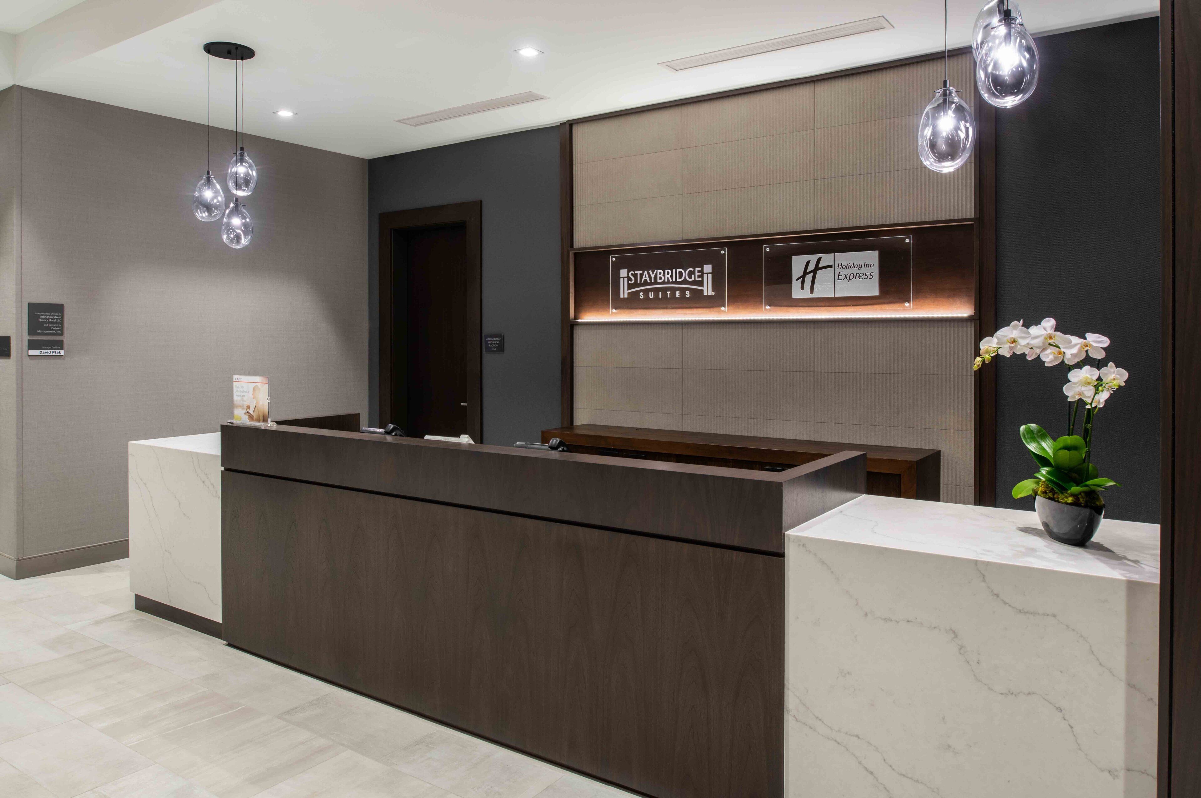 Un mostrador de recepción con acabado de madera oscura, un letrero de Staybridge Suites y un letrero de Holiday Inn Express. Hay una orquídea en maceta a la derecha.