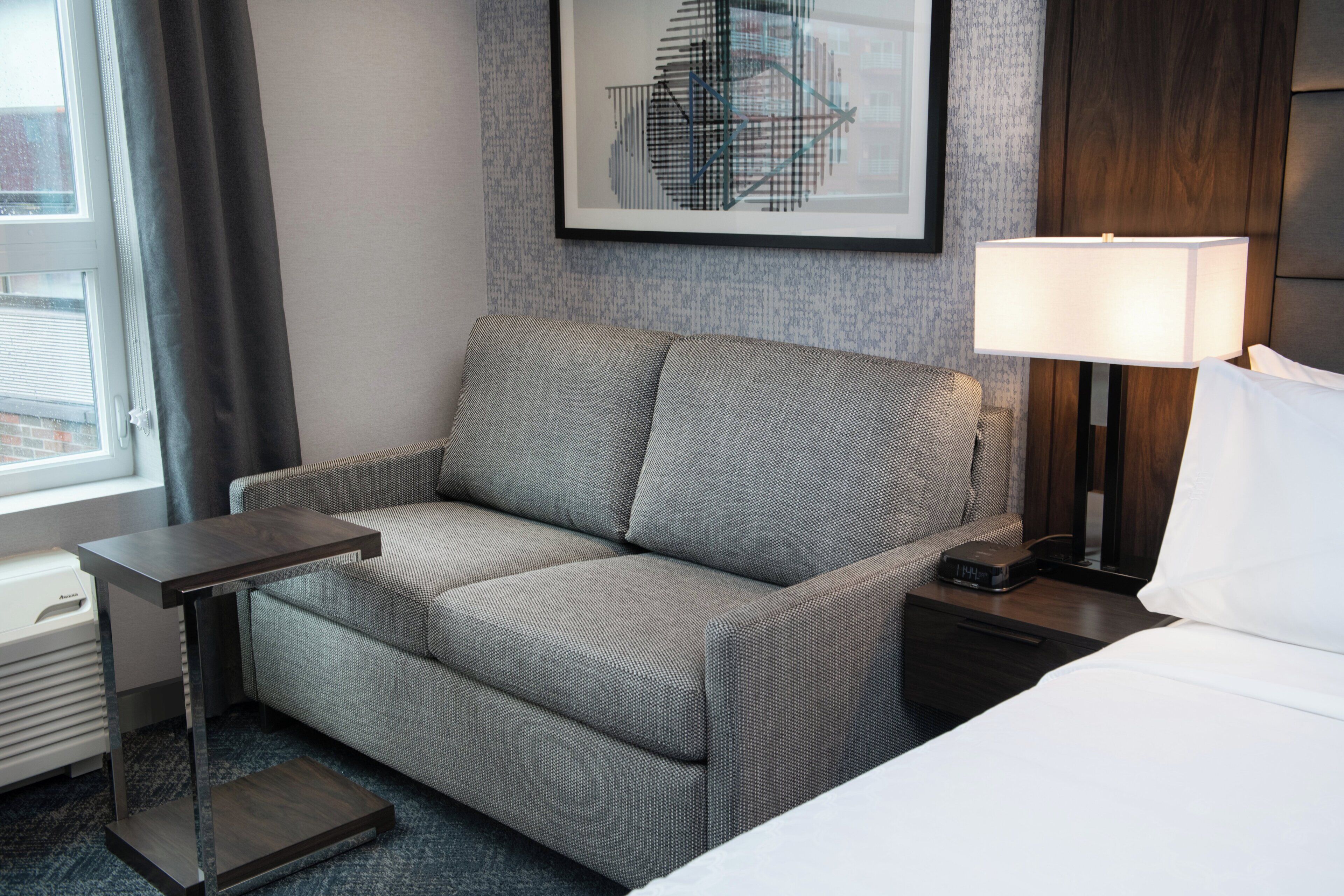 Una habitación de hotel con un sofá gris, una mesita, una lámpara y una cama.