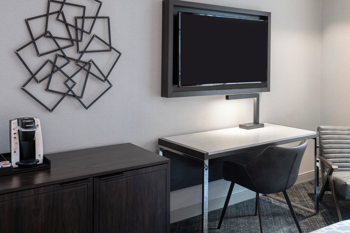 A modern hotel room with a flat-screen TV mounted on the wall, a desk with a chair, a coffee maker, and a decorative wall art piece.