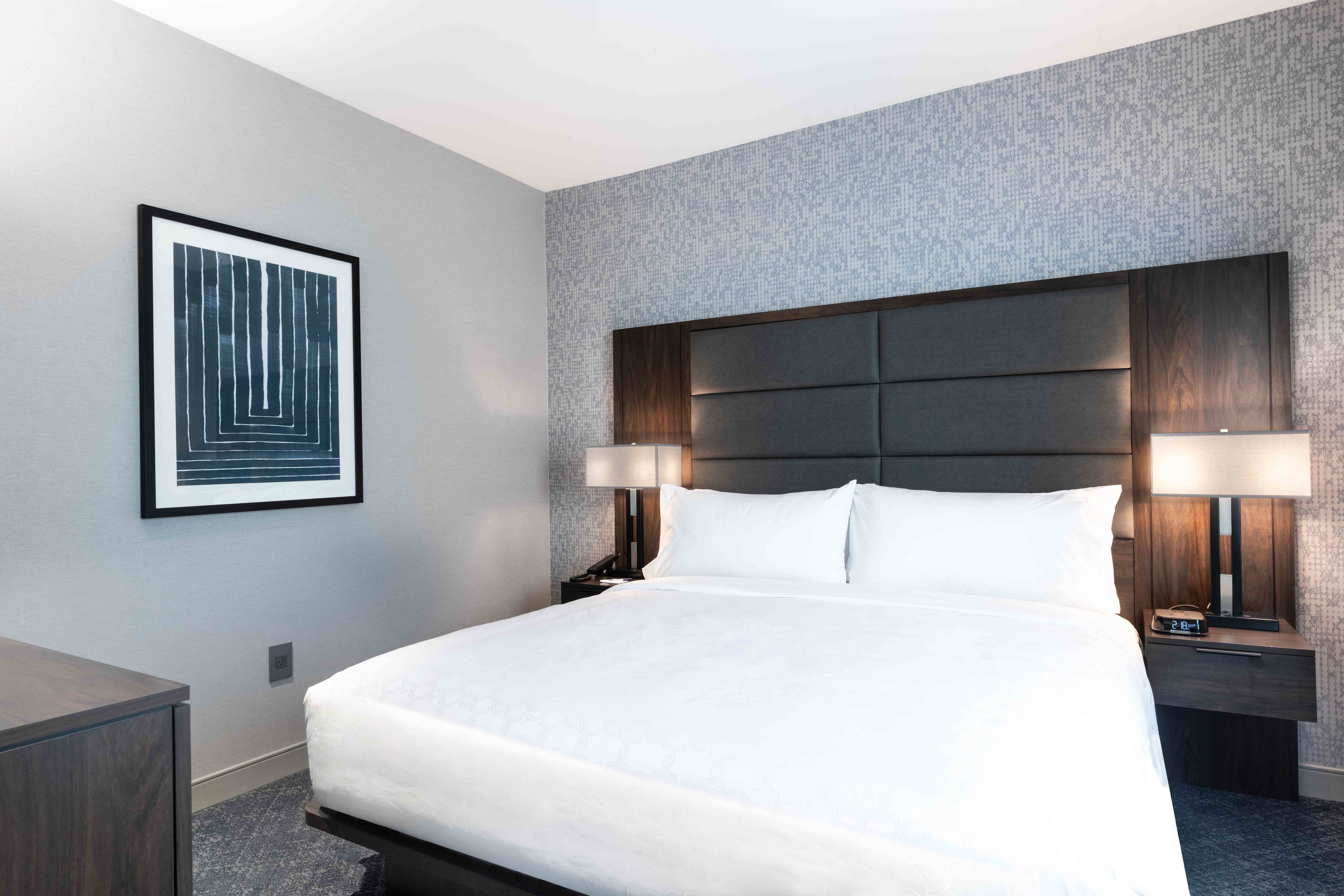 Habitación de hotel con una cama grande, dos mesitas de noche con lámparas, una obra de arte enmarcada en la pared y un revestimiento de pared texturizado.