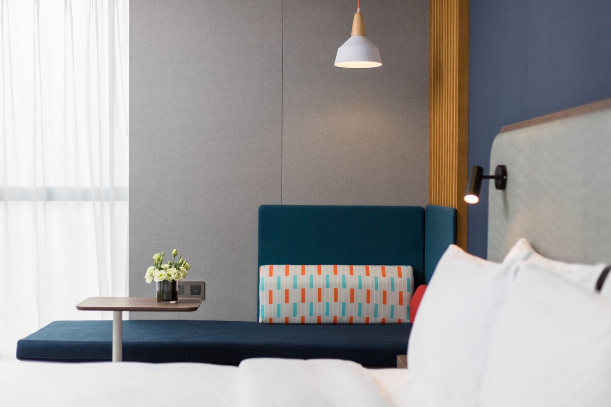 A modern hotel room with a bed, a small table with a vase of flowers, and a wall-mounted lamp.
