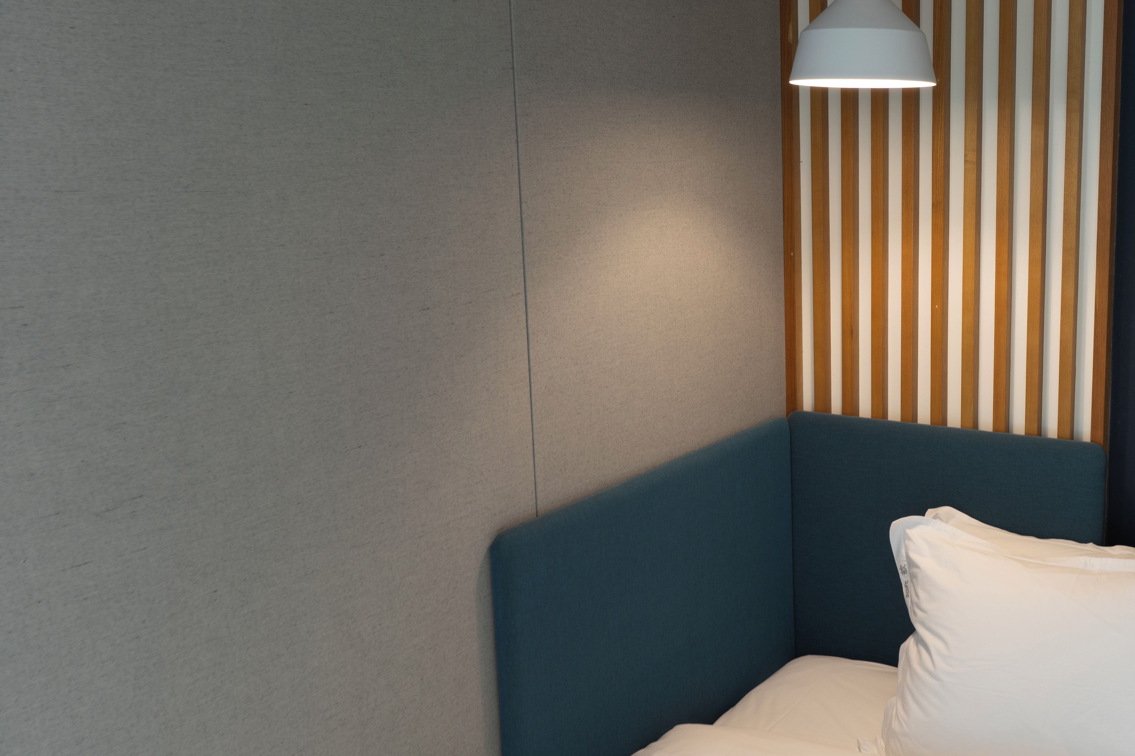 A corner of a room with a blue upholstered headboard, a white pillow, and a wall with vertical wooden slats.