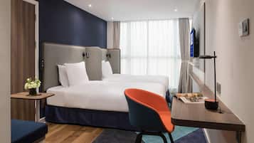 A modern hotel room with a large bed, a desk with a chair, a TV, and a window with curtains.