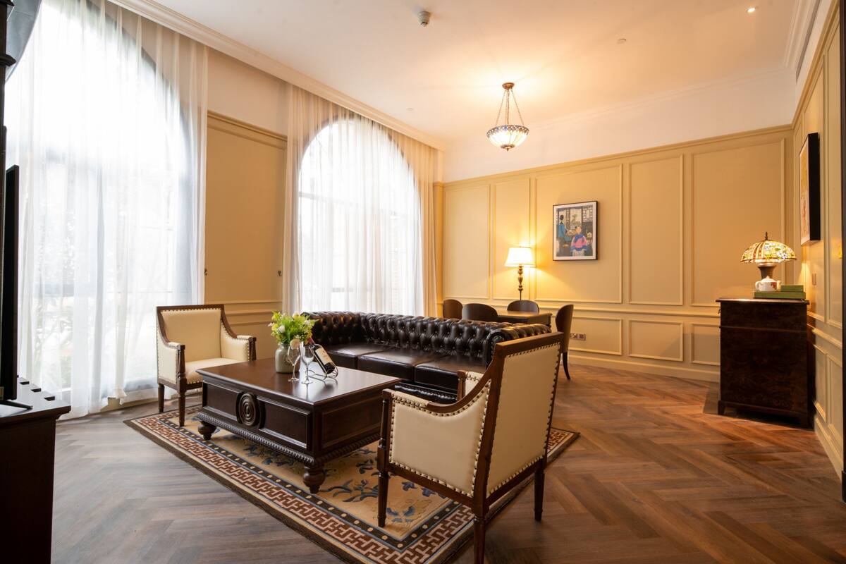 A room with a leather sofa, wooden coffee table, and a patterned rug. There is a chandelier, a lamp, and a framed picture on the wall.