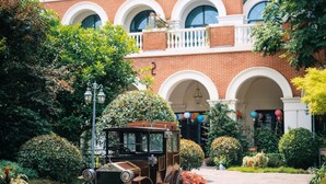 Une voiture ancienne est garée devant un bâtiment en briques, doté de fenêtres en arc de cercle et d’un balcon.