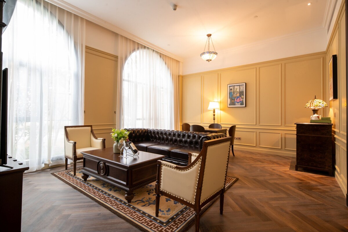 Een kamer met een leren bank, een houten salontafel en een kamerbreed tapijt. Er is een kroonluchter, een lamp en een lijst met een afbeelding aan de muur.