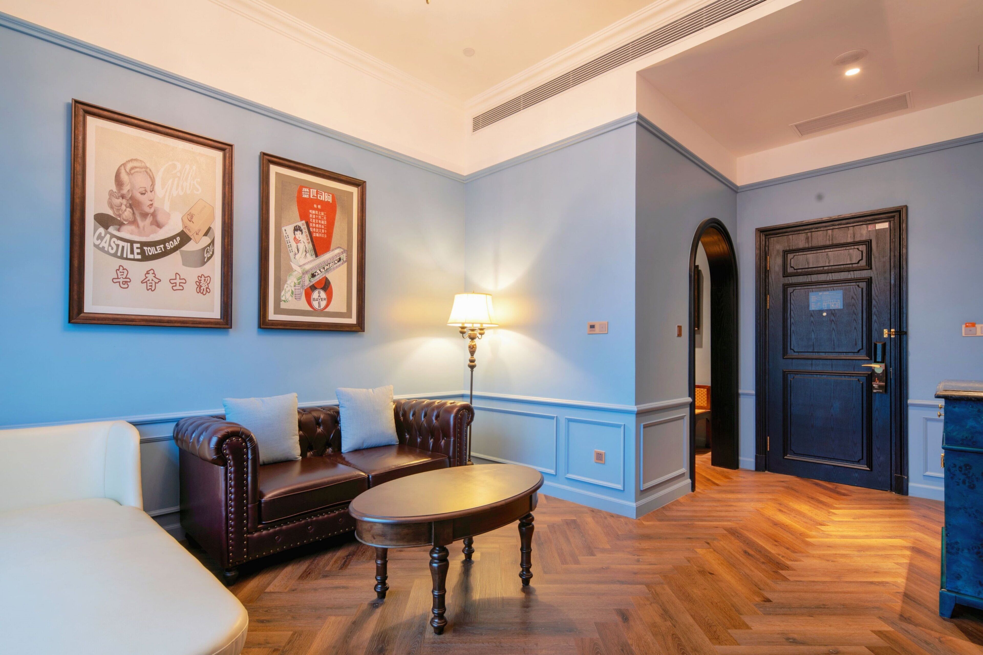 A room with a dark brown leather sofa, a round wooden table, and a blue wall with two framed advertisements.