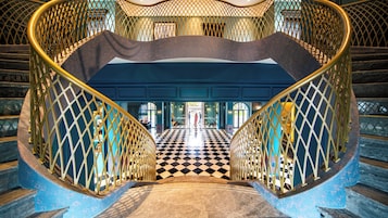 Una imponente escalera con piso a cuadros y ornamentales barandales dorados que conduce a una habitación con paredes azules y un amplio ventanal.