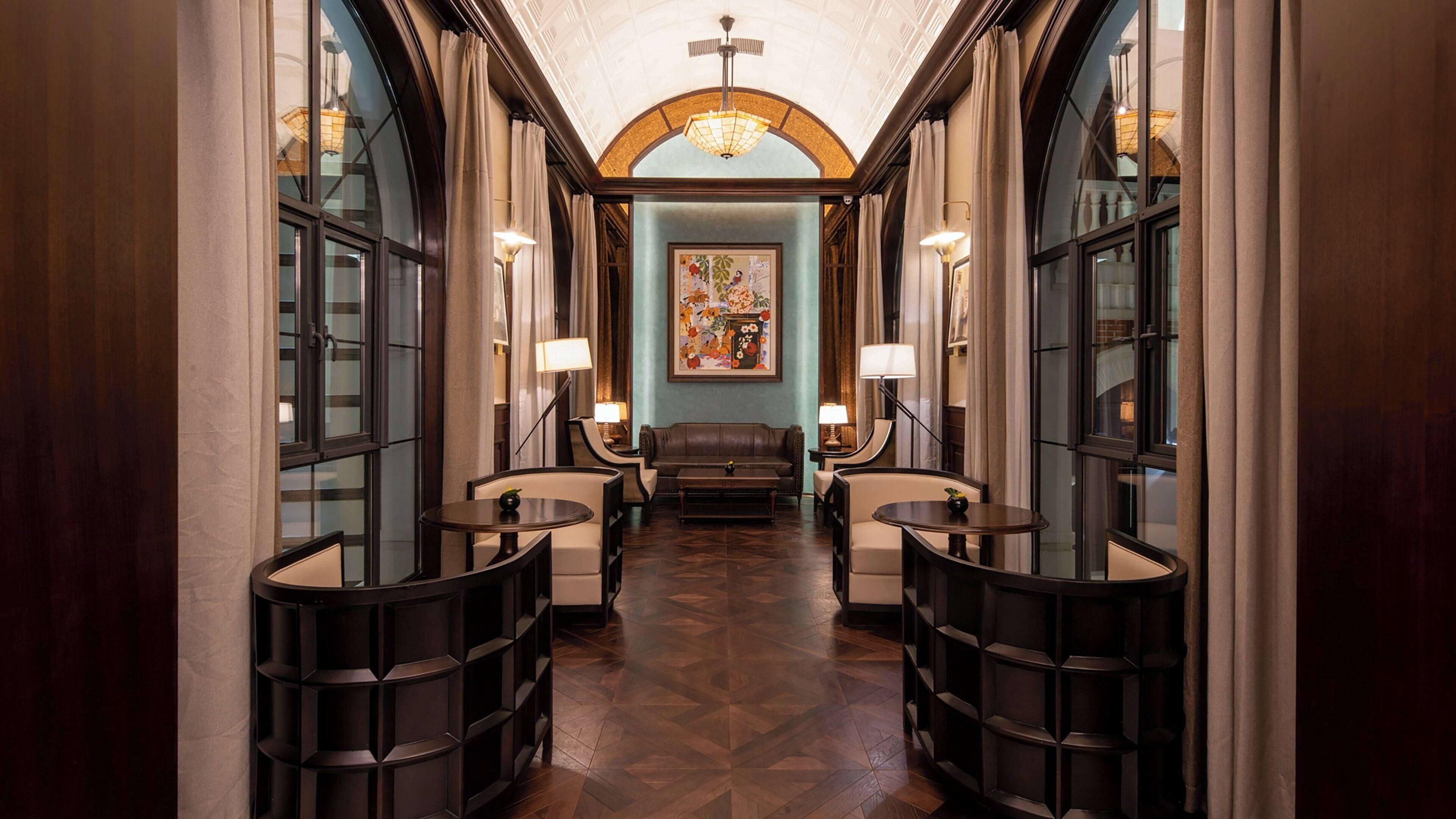 A hallway with a central seating area, flanked by two tables with lamps, leading to a room with a large painting.