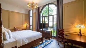 Une chambre spacieuse avec un grand lit, un bureau et une chaise, un lustre et une grande fenêtre donnant sur un paysage verdoyant.