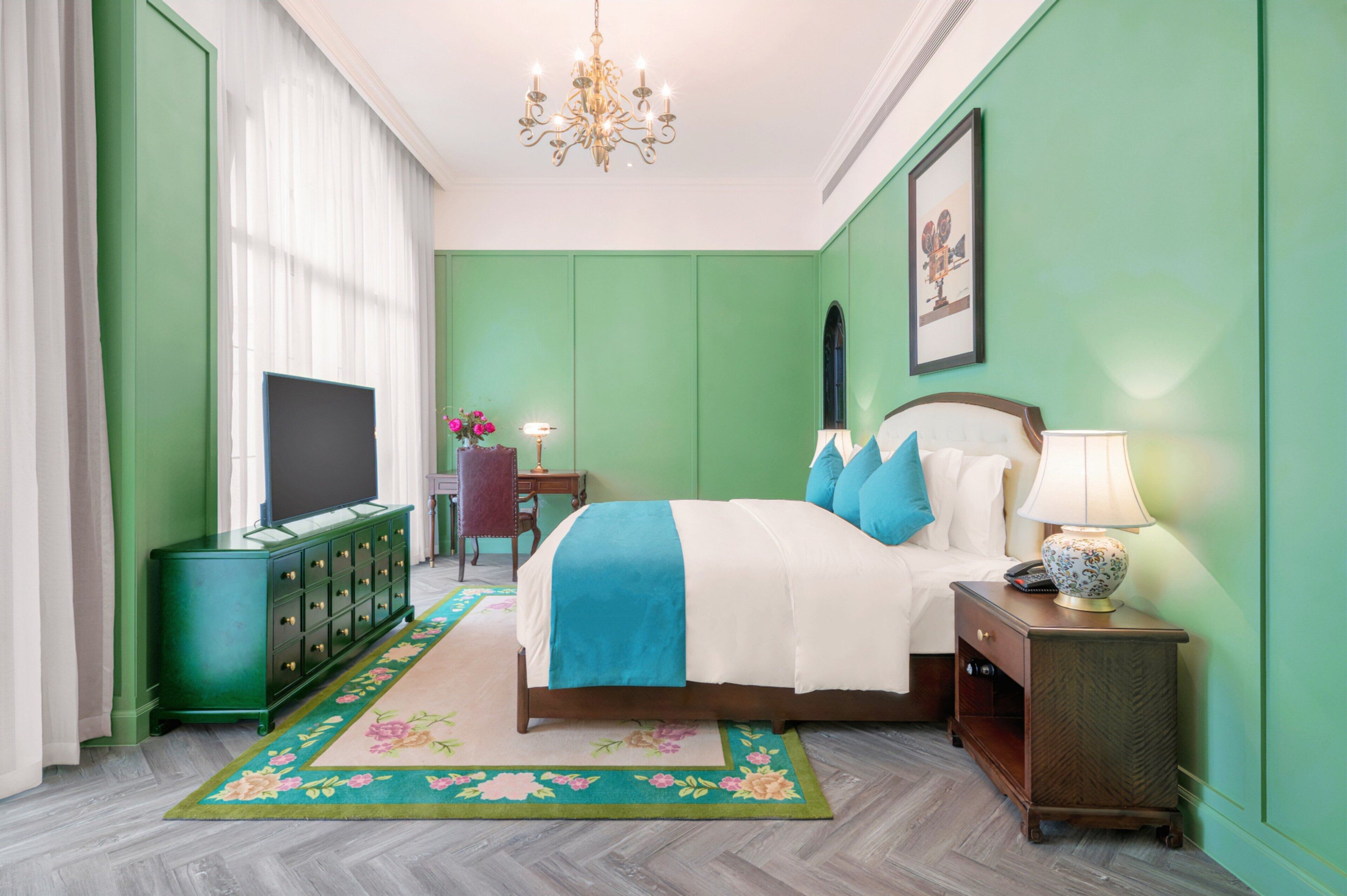 A hotel room with a bed, a television, a dresser, a nightstand, and a chandelier.