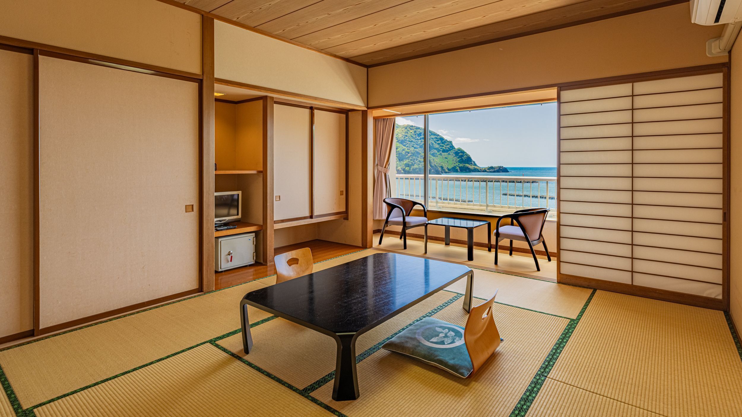Una habitación con suelo de tatami, una mesita baja, sillas, un televisor y vista al mar.