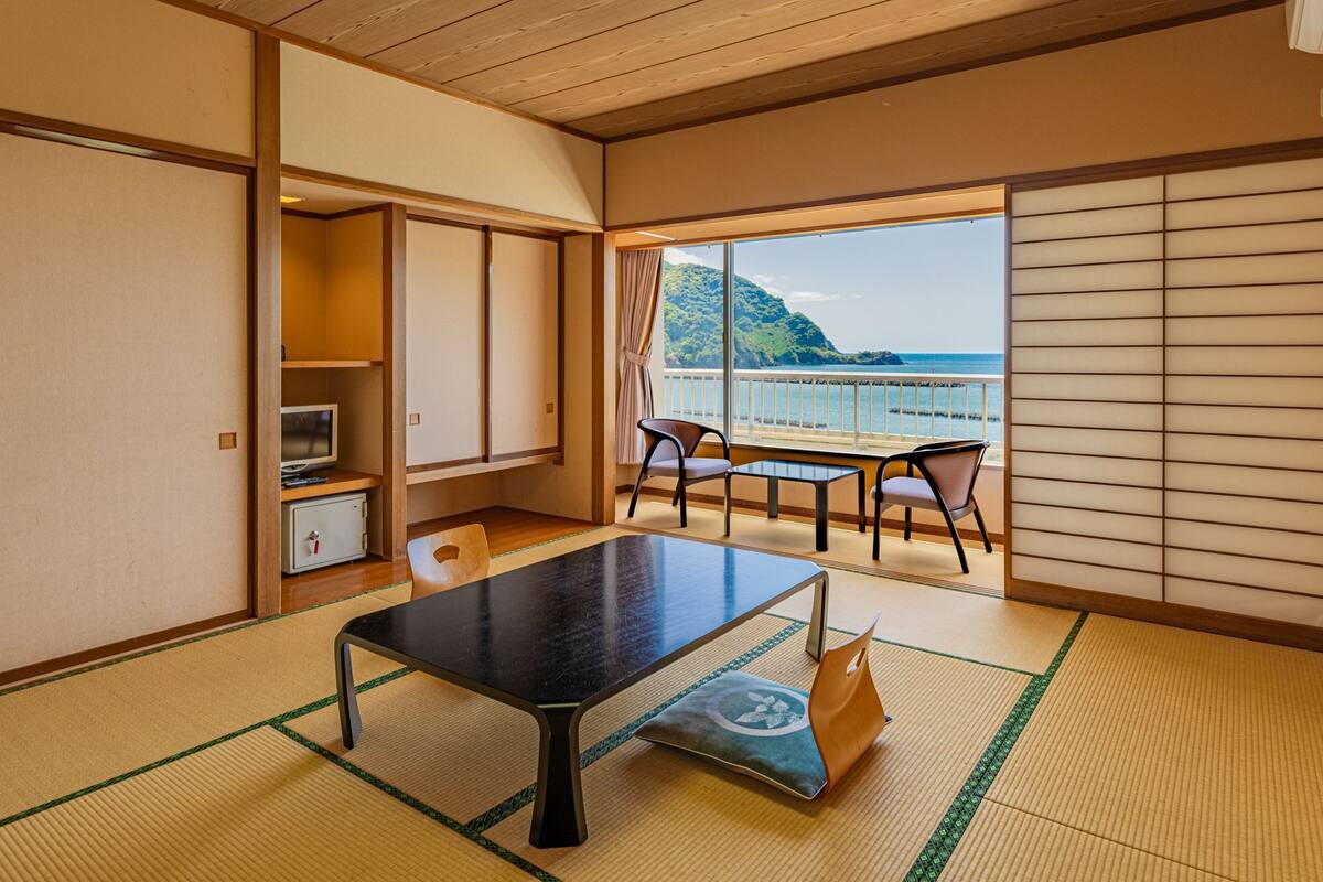 Een kamer met tatami vloer, een lage tafel, stoelen, een televisie en uitzicht op zee.