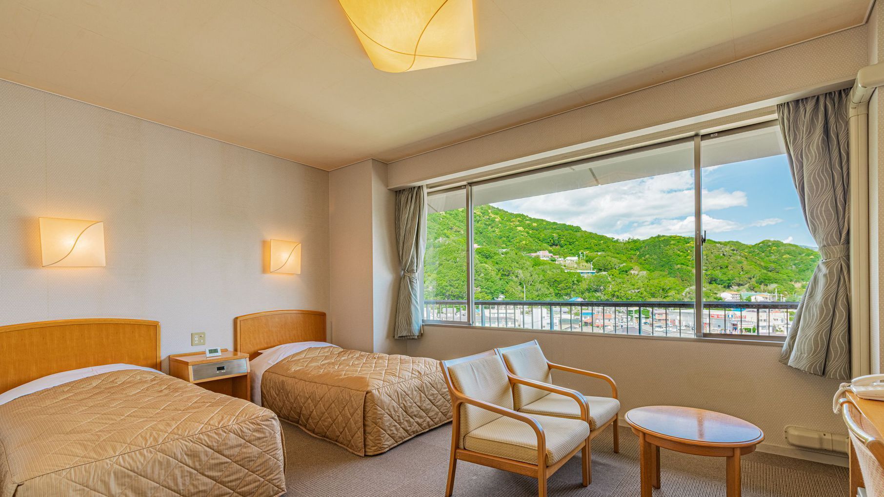 Habitación de hotel con dos camas, una silla, una mesita y un ventanal con vistas a un paisaje verde.