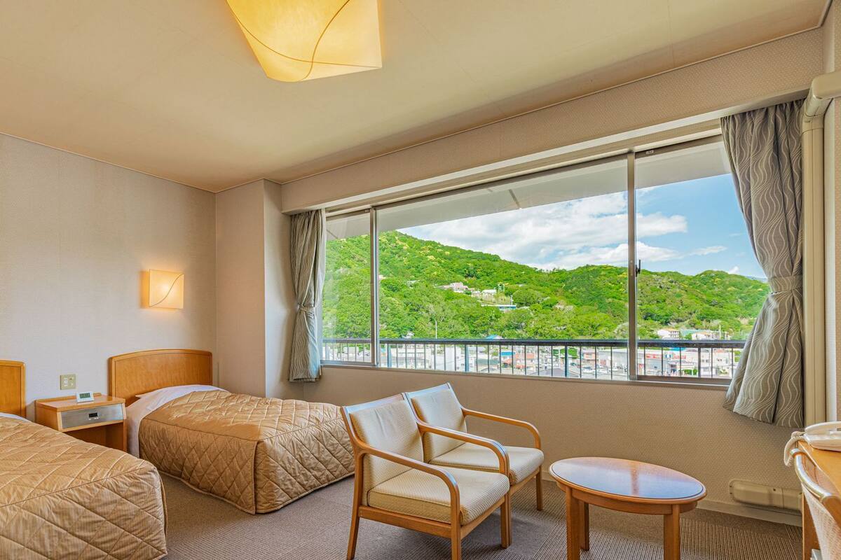 Een hotelkamer met twee bedden, een stoel, een tafeltje en een groot raam met uitzicht op een groen landschap.