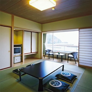 Een kamer met tatami vloerbedekking, een lage tafel, stoelen, een televisie en uitzicht op een waterpartij.