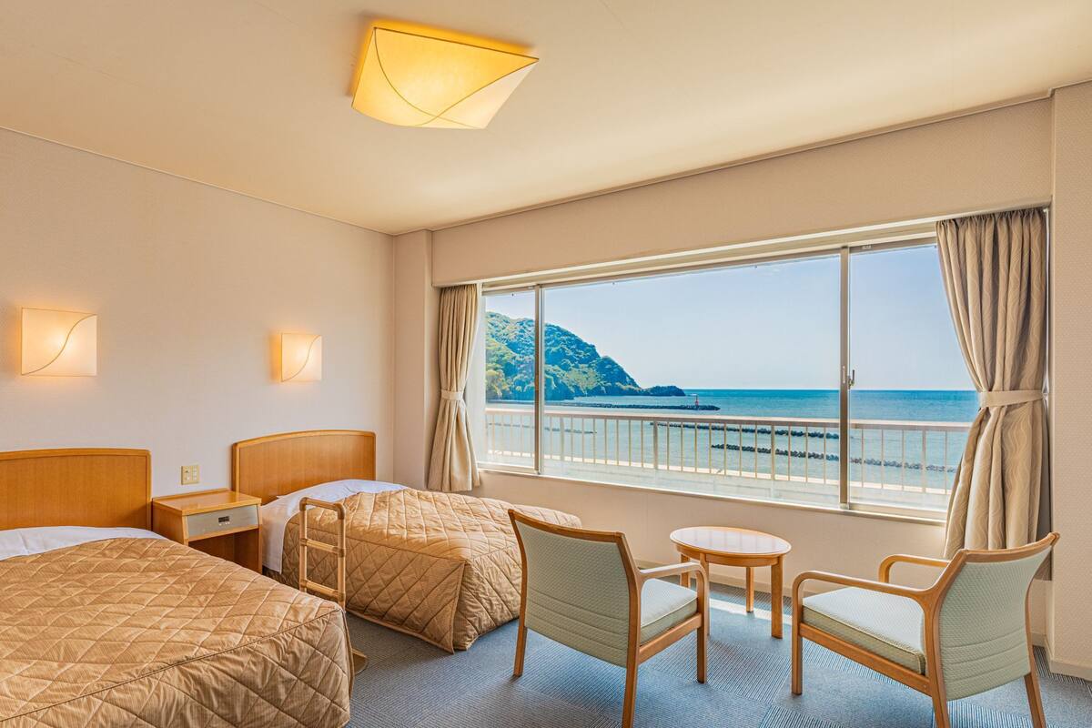 Hotelkamer met twee bedden, een tafeltje, twee stoelen, een nachtkastje en een groot raam met uitzicht op zee en een klif.