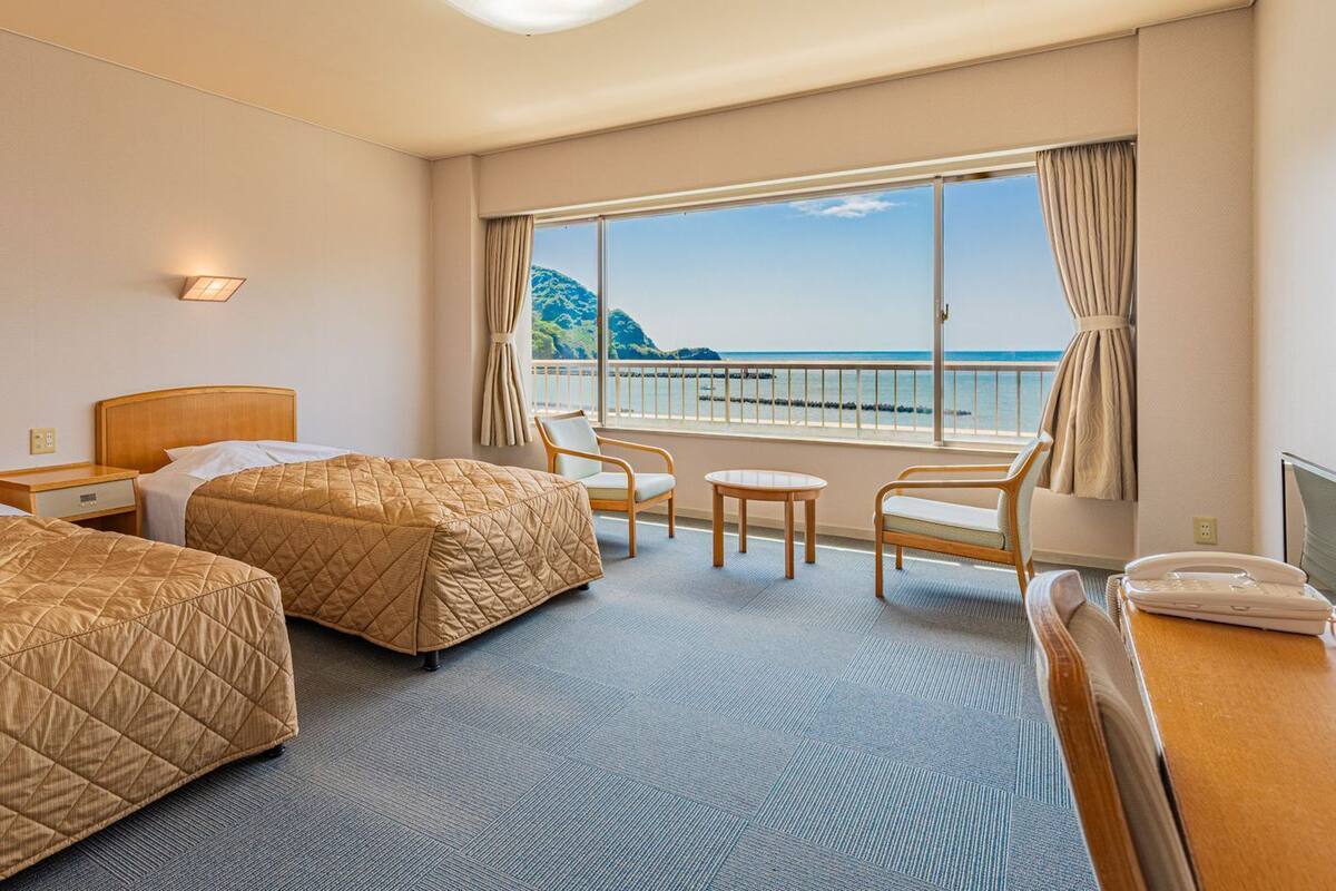 Een hotelkamer met twee bedden, een kleine tafel, een stoel, een bureau en uitzicht op zee.