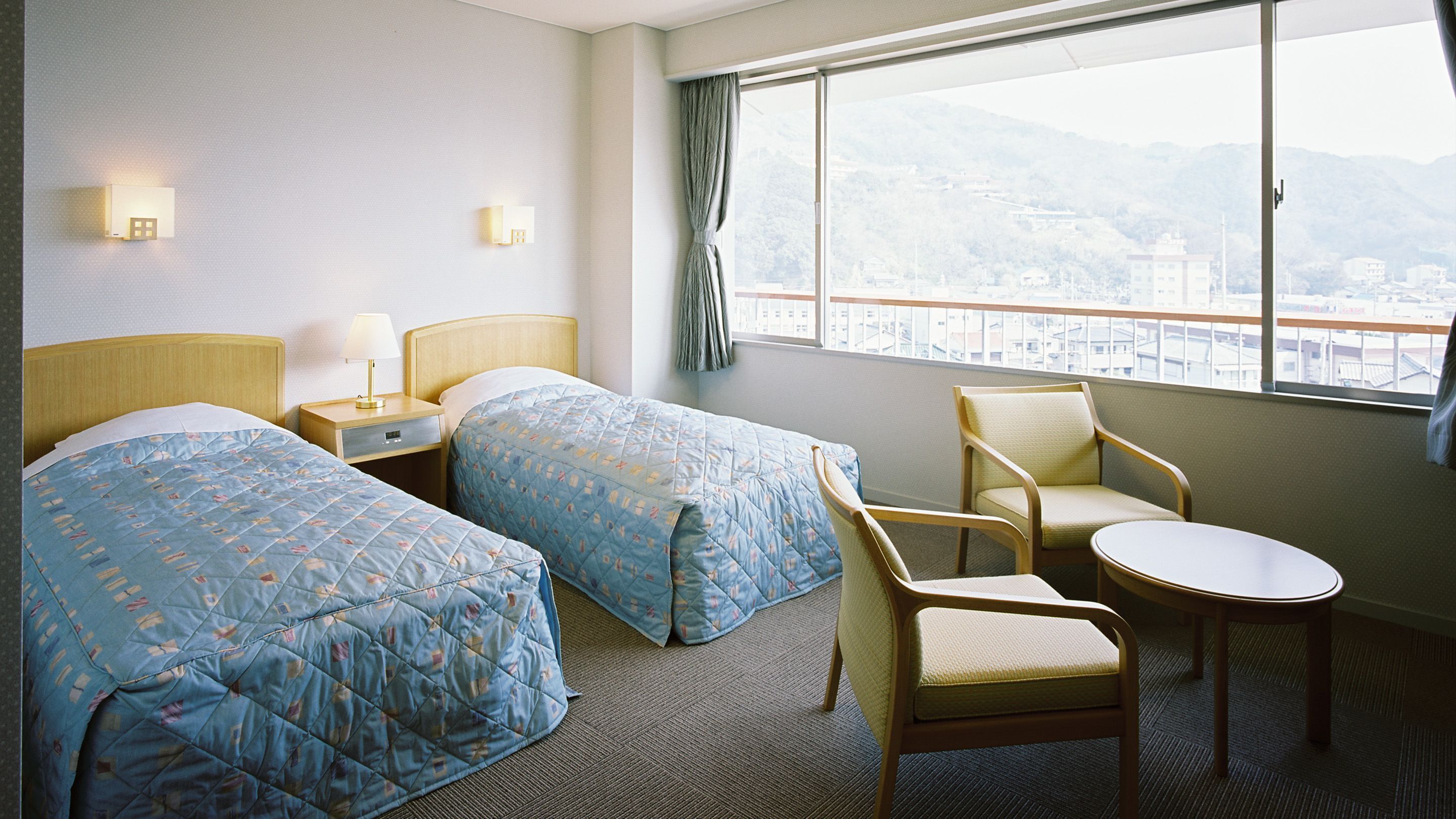 Habitación de hotel con dos camas, una mesita de noche, una silla, una pequeña mesa y una ventana con vistas a la ciudad.