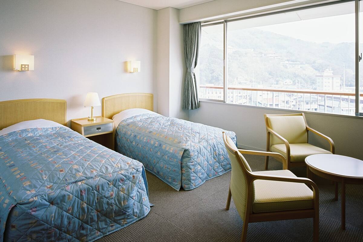 Een hotelkamer met twee bedden, een nachtkastje, een stoel, een klein tafeltje en een raam met uitzicht op een stadsbeeld.
