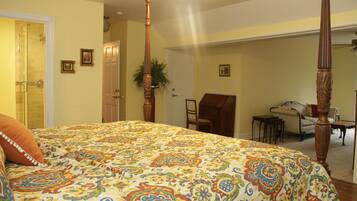 A bedroom with a large bed, a wooden four-poster headboard, a chair, a small table, and a door leading to another room.