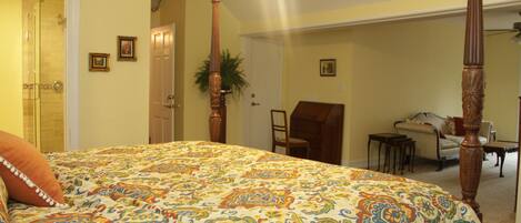A bedroom with a large bed, a wooden four-poster headboard, a chair, a small table, and a door leading to another room.